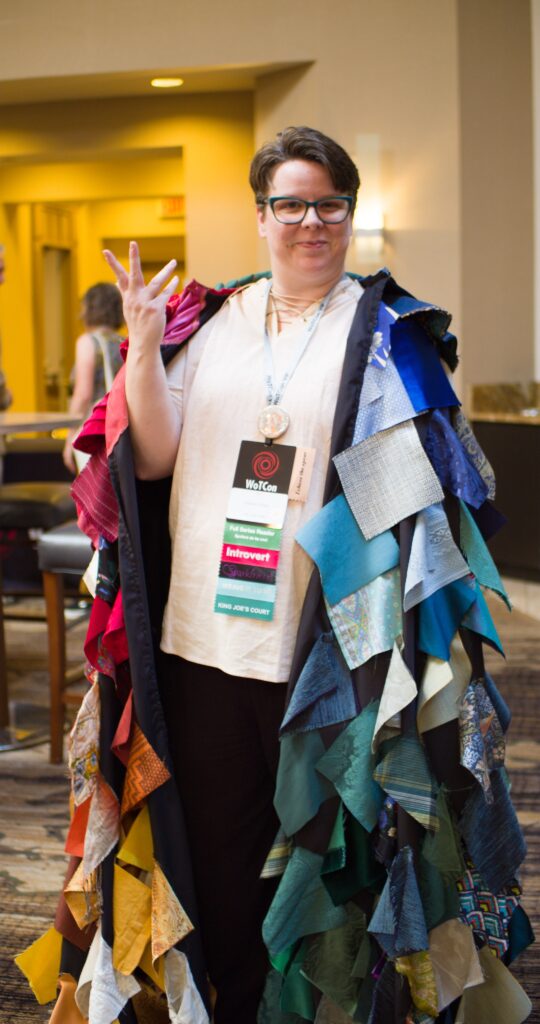 Eilonwy in colorful gleeman’s cloak with multicolored and textured patches. They have short brownish hair, pale skin and glasses.