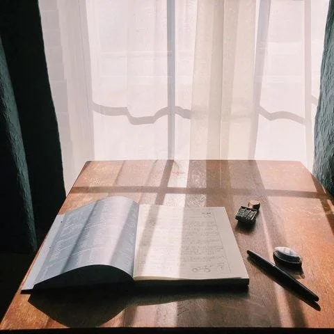 Blue notebook on a wooden table with light filtering through sheer curtains. A pen and date stamp sit nearby.
