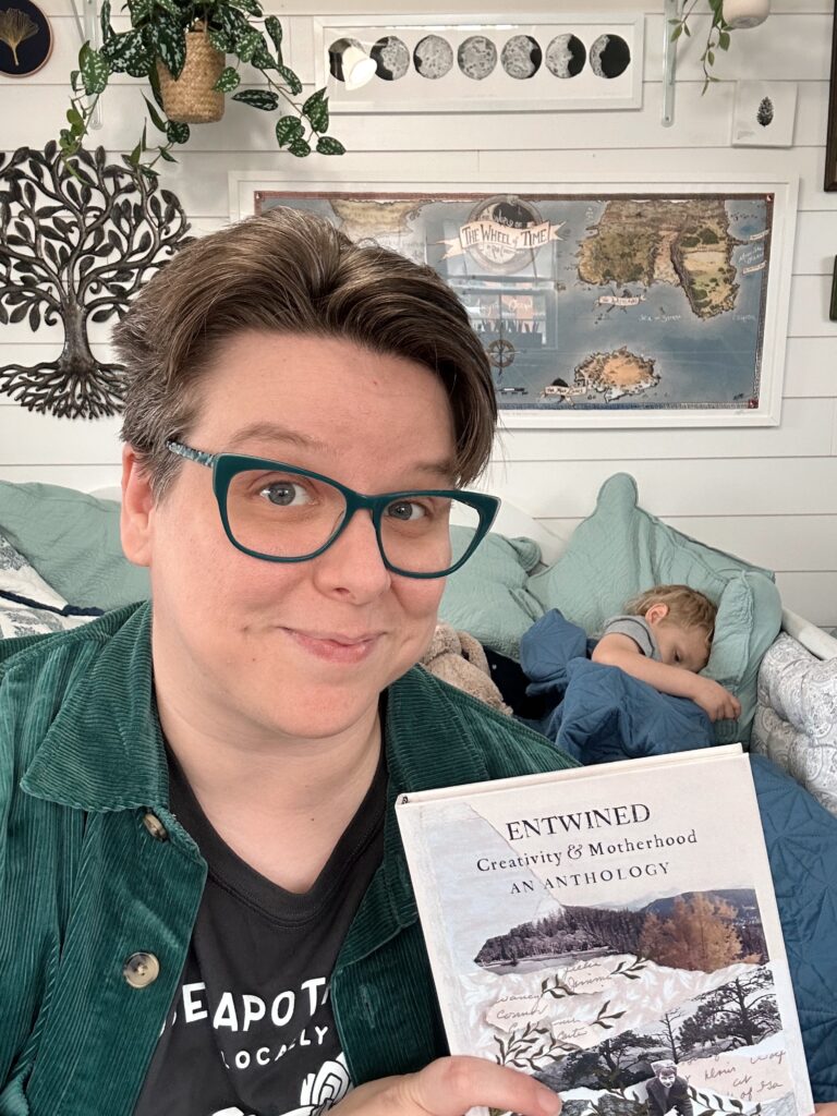 Selfie holding a copy of Entwined in my studio with my kid playing video games on the daybed behind me. I have on a Rose Apothecary shirt, green corduroy overshirt, and green glasses. I have short brownish hair and blue eyes.