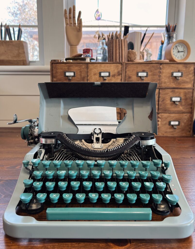 Teal green typewriter with an open carriage for cleaning sits on a wooden desk. Library style drawers and wart supplies are behind.