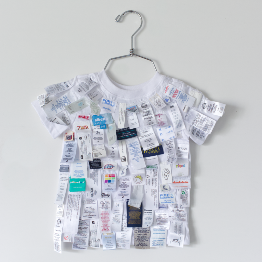 White child sized t shirt on a silver clothes hanger covered in clothing tags of various brands and sizes.