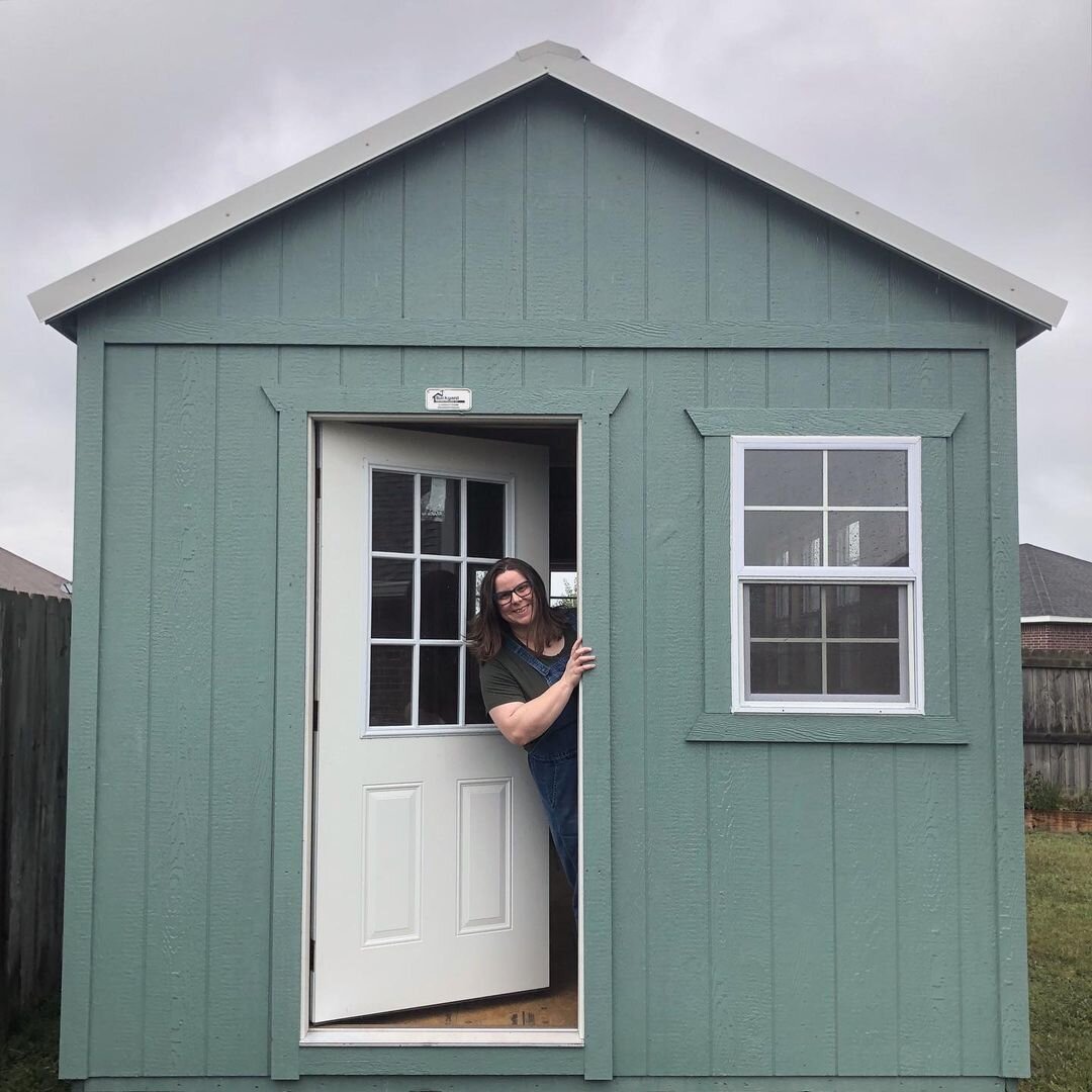 I am peeking out of a gray green studio shed while wearing overalls.