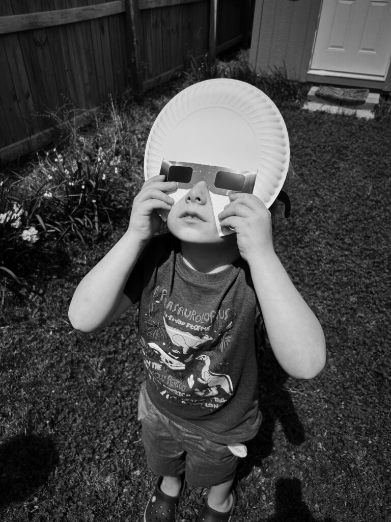 Black and white photograph of a child looking up through eclipse glasses slotted through a paper plate