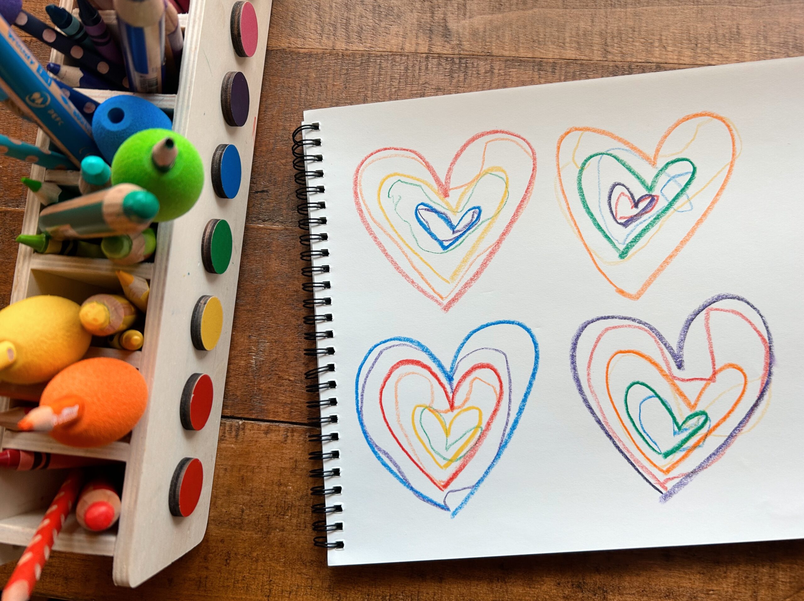 Nested rainbow hearts drawn with crayons. Rainbow sorted colored pencils and art supplies to the left.