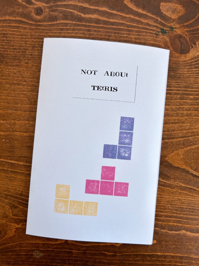 Not About TETRIS zine on a wooden table. The title is letterpress printed and three printed blocks in purple pink and yellow are arranged as if to pile up.