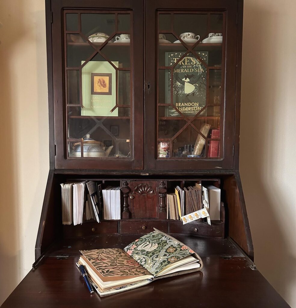 Antique secretary with glass doors covering shelves displaying Anne of Green Gables and Tress of the Emerald Sea. Fold out desk has pigeon holes with envelopes and a traveler's notebook with William Morris inserts sits on the desk.