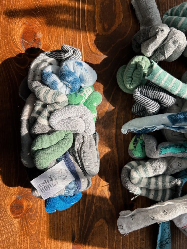 Soft sculpture of a brain made from baby clothes. One hemisphere is sewn from baby socks, onesies and washclothes. Scraps sit on a wooden table to the side.