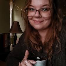 Photo of Jessica Allowski wearing glasses and holding a coffee cup with fairy lights behind