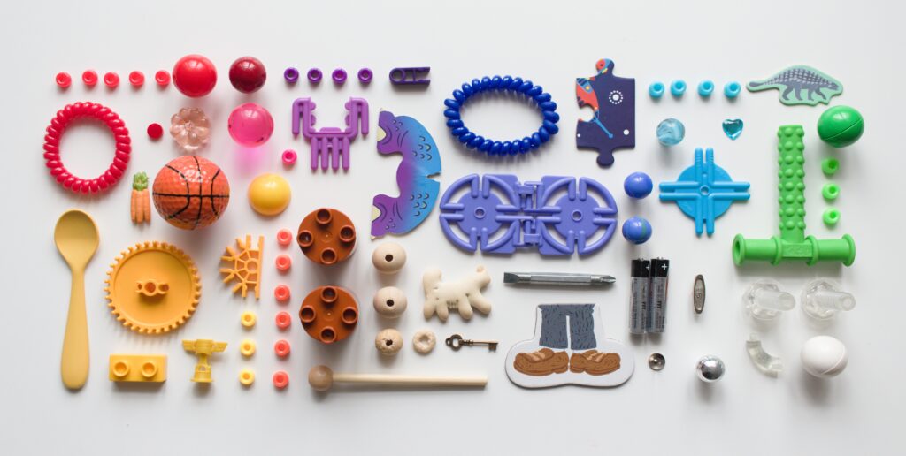 Small toys arranged by color including red beads, marbles, bracelet, orange golf ball, carrots, beads, yellow spoon, cog, LEGO DUPLO, trophy, beads, K'nex, brown round DUPLO, wooden beads, drumstick, purple plastic shapes, k'nex, and beads.