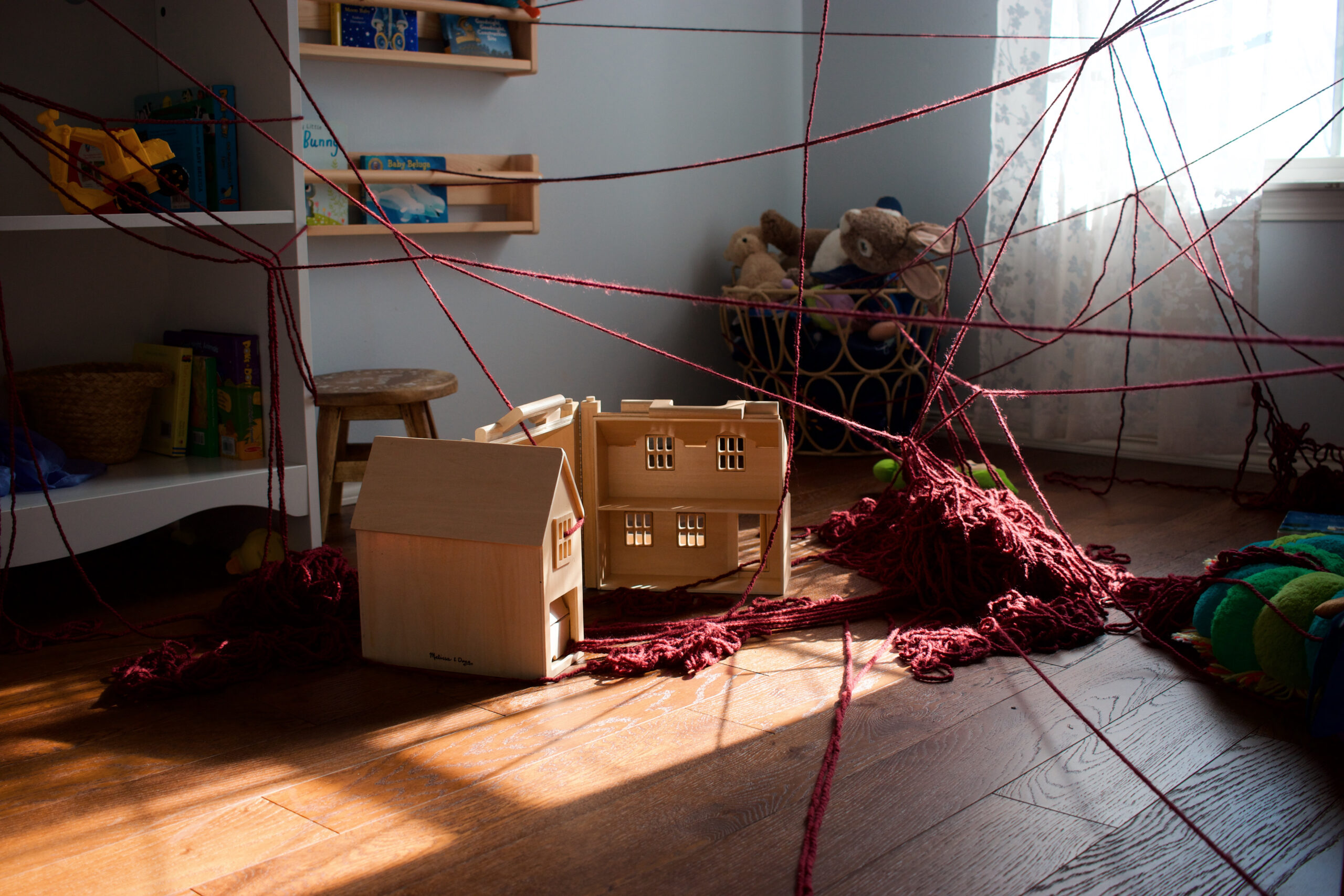 Still photograph of temporary installation in my child's nursery. Dark red yarn is strung around the room. Sunlight throws shadows across yarn and wooden dollhouse.