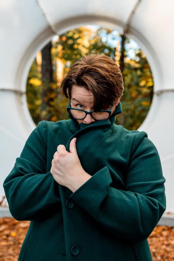 Sarah Shotts hides face in dark green coat and peeks over green glasses. Their brownish hair falls into their face. Buckminster Fuller's Fly Eye Dome frames their face from behind.