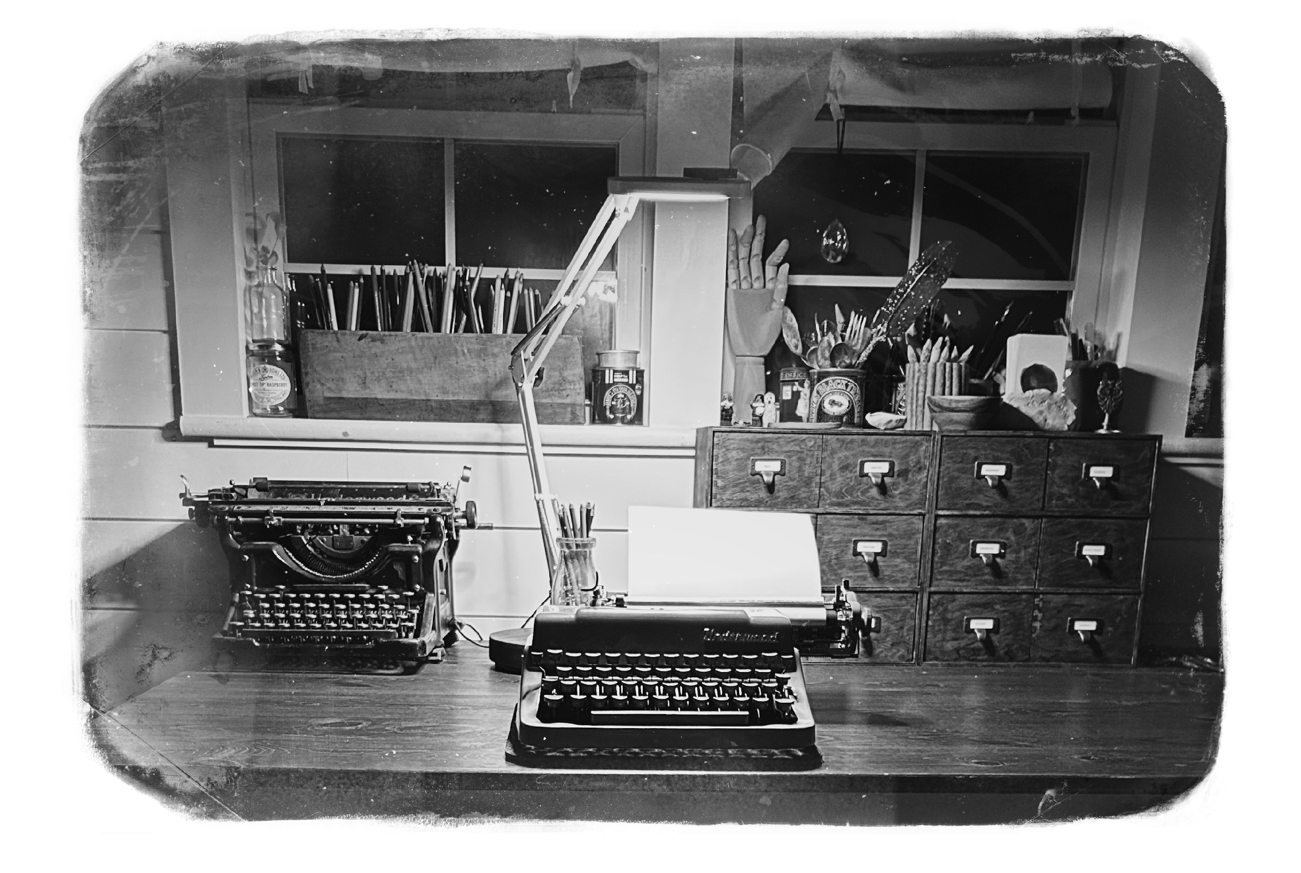 Black and white photograph of writing desk at night with two vintage typewriters and a set of library style drawers in the light of a a lamp. Torn bottom edge.