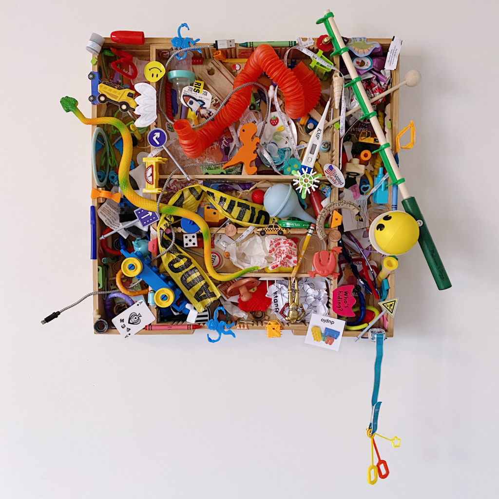 A visual cacophony of broken toys and the detritus of motherhood fill a bamboo cutlery tray mounted on the wall. Most easily visible are a plastic snake, foam Pac Man, fishing pole, red tube, DUPLO train, caution tape, and bubble wands. There are also various candy wrappers, stickers, crayons, small toys, and broken objects throughout.