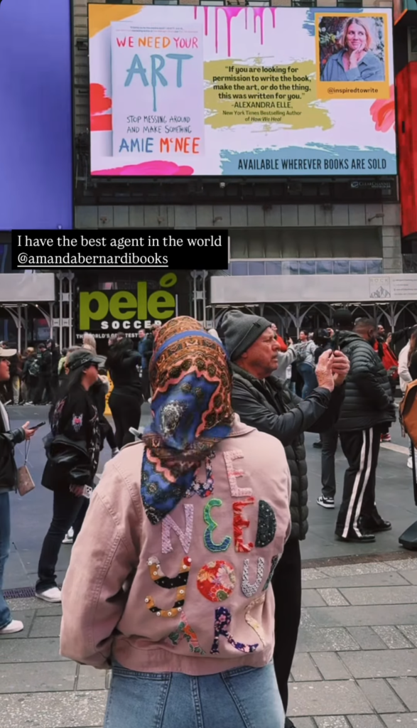 Amie wears a custom jacket with We need your art quilted on the back and looks at her book on a sign in Times Square.