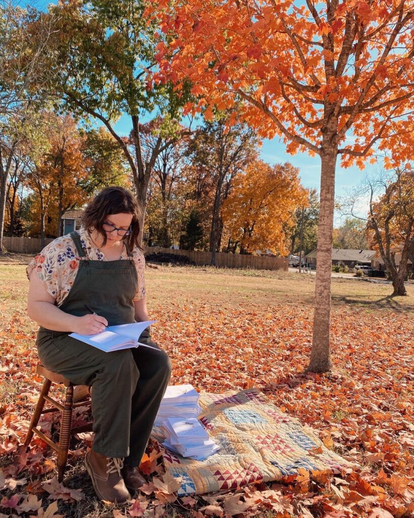 I am wearing overalls singing books. I'm sitting under a maple tree next to a pile of books on a quilt.