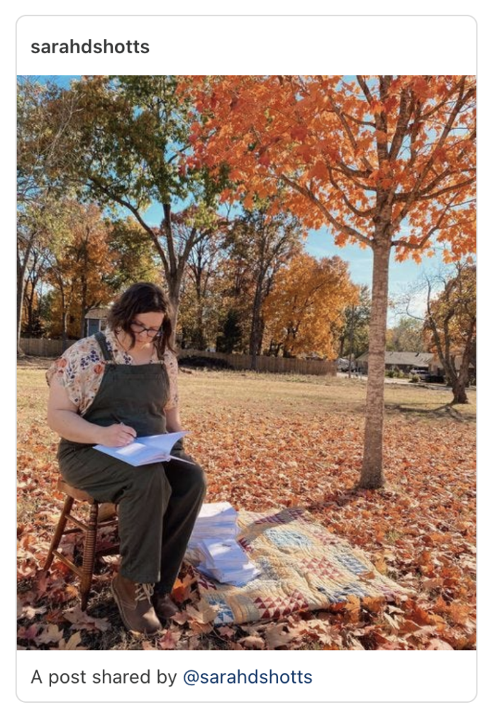 I am sitting under an orange maple tree next to a pile of books on a quilt signing books.