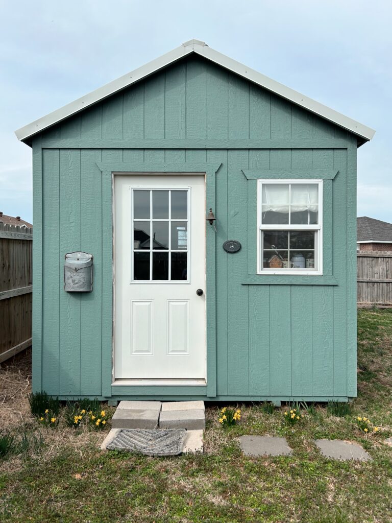 Gray green studio with white door and gray metal old fashioned mailbox. A row of tiny daffoils grown in front of a path of square stepping stones. The grass is mostly dormant.