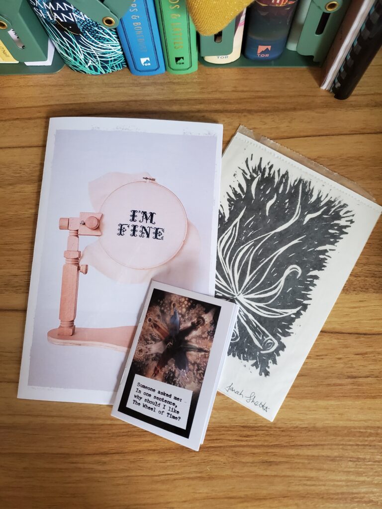 My I'm Fine zine, Wheel of Time mini zine and a campfire linocut print lay on a table by a row of blue and green books.