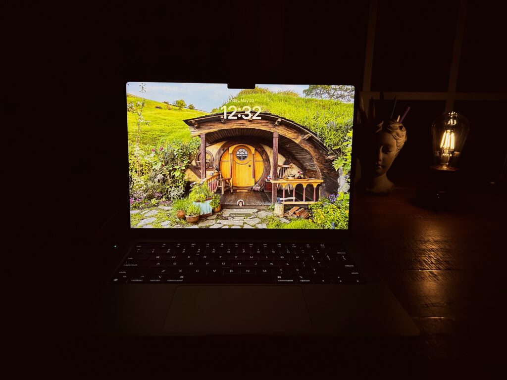Laptop with yellow hobbit hole wallpaper. An edison bulb lamp sheds a dim golden glow on a goddess vase filled with pens. The workspace is otherwise shrouded in darkness.