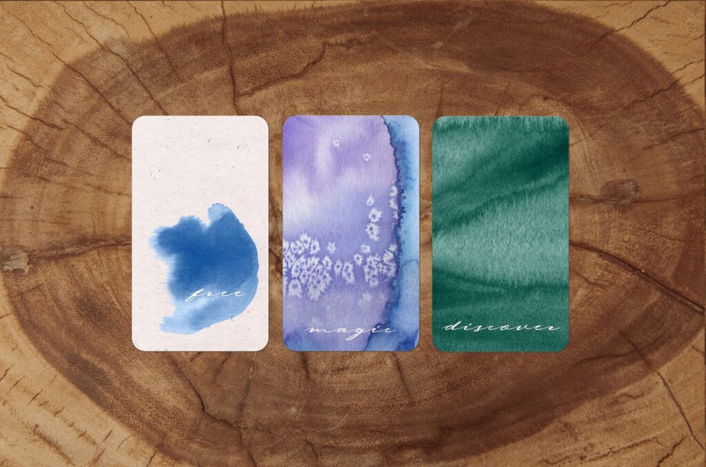 Three cards on a tree trunk background. They each have words in a white script overlaid a watercolor wash. The first is free over a light blue wash that evokes clouds. The second card is covered in purple and a bit of blue with salt sprinkled over to make textured sparkles. It is magic. The third is a teal blue wash covering the card and says discover.