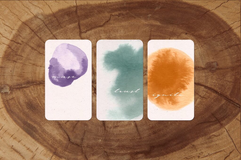 Three cards on a tree trunk background. They each have words in a white script overlaid a watercolor wash. The first is an irregular watercolor wash in purple and says muse. The second is a pale green that says trust. The third is a golden circle with texture evoking rays that says igite.