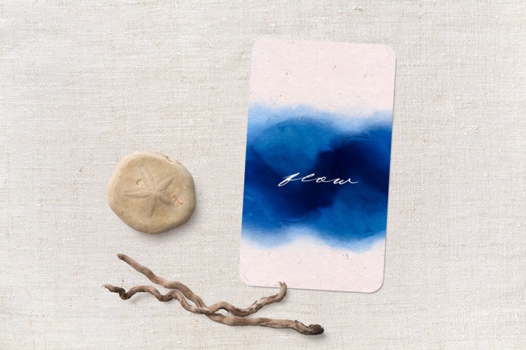 A mockup of a card with a seashell and driftwood. The card says flow and has overlaid watercolor washes in blue that evoke water.