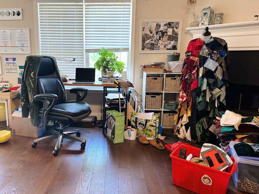 Laptop on desk in the living room surrounded by homeschool materials, dressing up box, shelves, boxes, clutter, and a gleeman's cloak on a dress form.