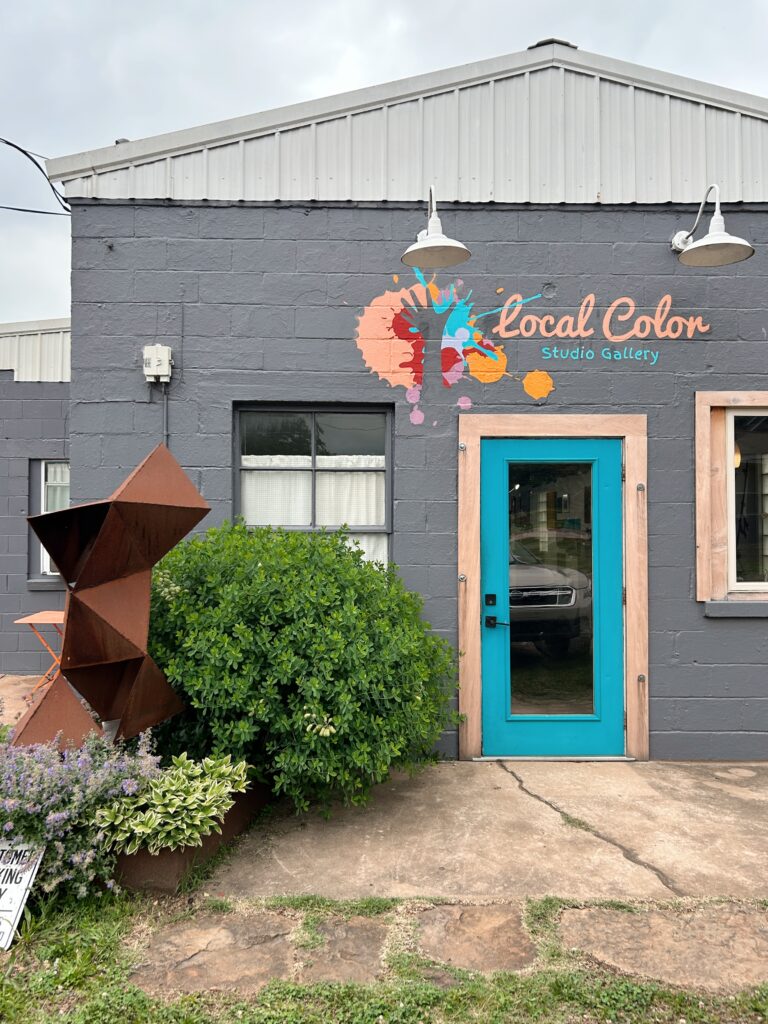 Local Color Studio Gallery a gray and white building with a colorful paint spattered logo and a bright turquoise door. A large baptisia plant grows to the side and a large metal sculpture of triangles stacked in both positive and negative space.