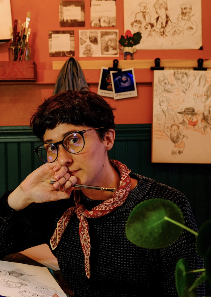 Gracie has short dark hair and glasses and is wearing a bandana and holding a paintbrush.