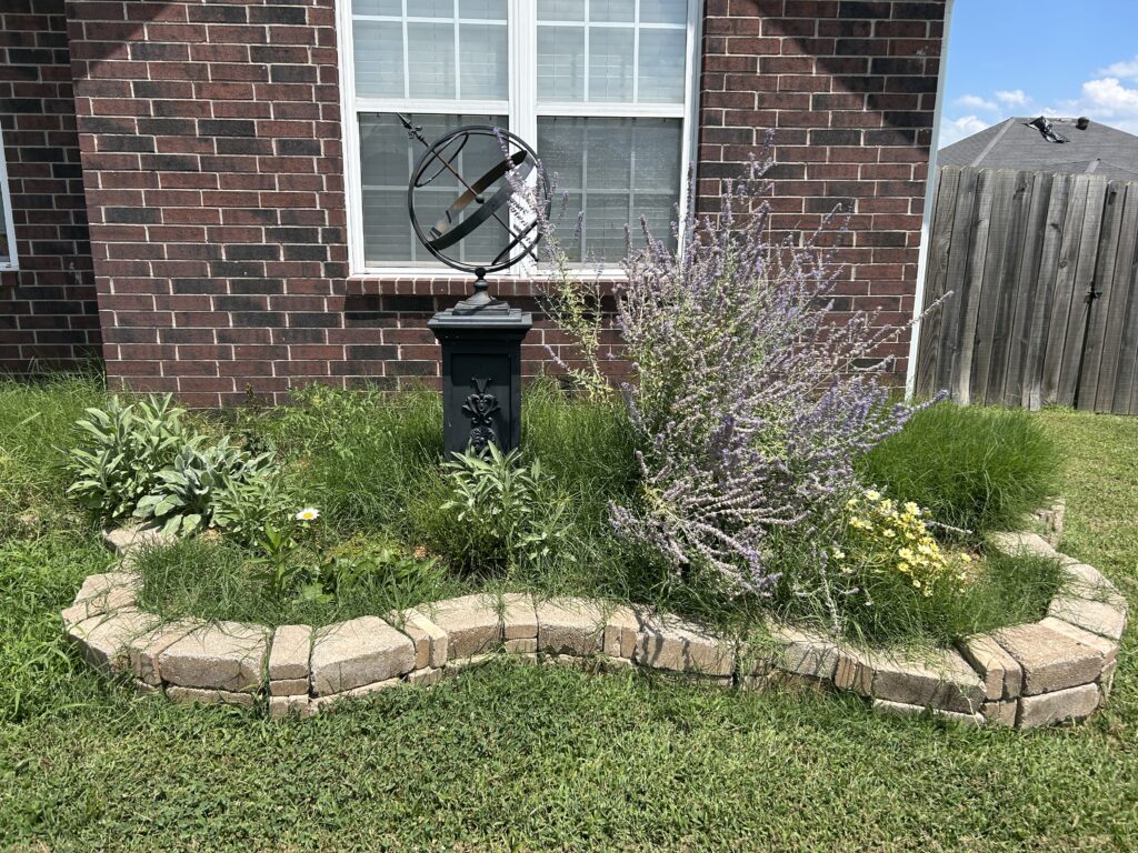 Brick house with an undulating garden bed overrun with grass. A black armillary sphere and purple russian sage pierce the weeds.