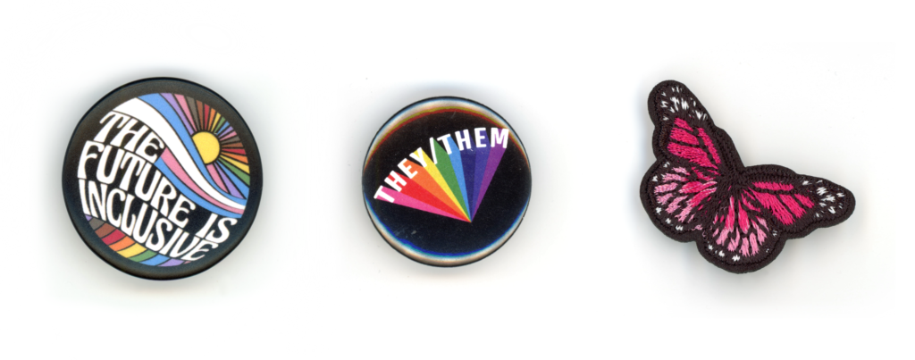 Two round pins and one pink butterfly patch. The first pin says, "The future is inclusive" and shows a wave and sunrise in pride flag colors. The second says they/them and a flare of rainbow coming off the letters.