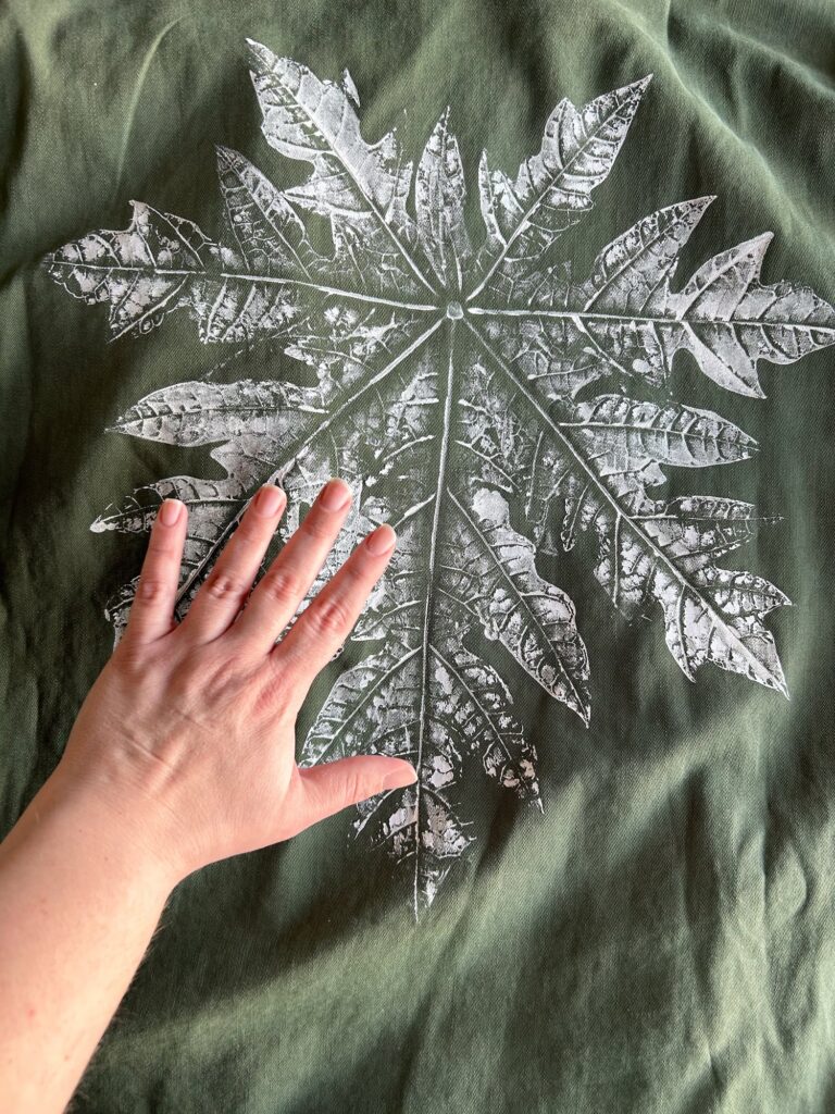 Giant papaya leaf printed with white paint on the back of a green jacket with my hand for scale.  It is roughly 4 times the size of my hand.