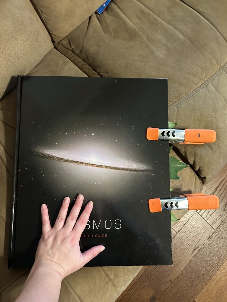 Giant Cosmos book clamped shut with my hand for scale. Edges of the papaya leaf peek out the edge.