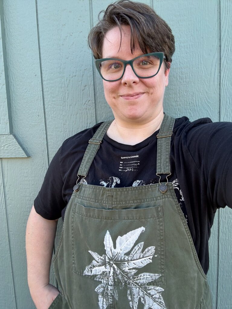 I am taking a selfie in my green overalls with white papaya leaf on the front. I am smiling and wearing green glasses. I have short brownish hair and pale skin.