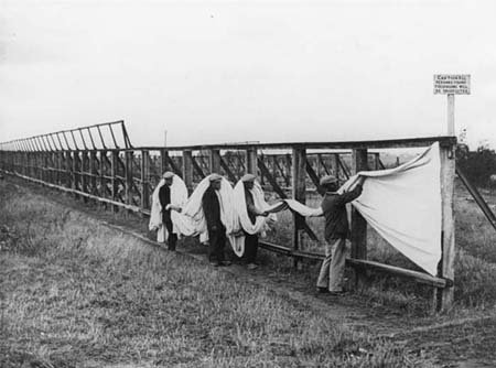 Black and white photograph of figures removing a large swathe of white cloth from a wooden frame with tenterhooks