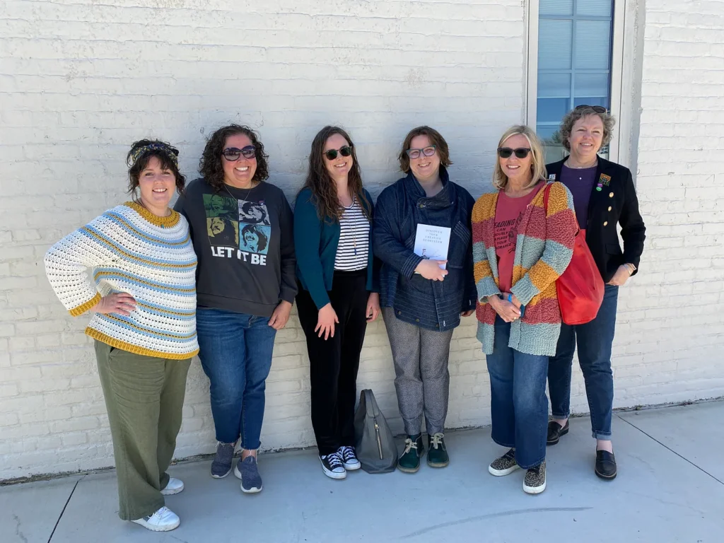 Five women and a nonbinary author standing against a white brick wall smiling. It's chilly weather so we are all in various sweaters and jean jackets. Three are wearing sunglasses. I am holding my book.