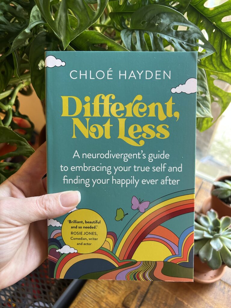 Different not Less by Chloe Hayden. Teal book with rainbows and butterflies held in a pale hand. House plants peek out behind.