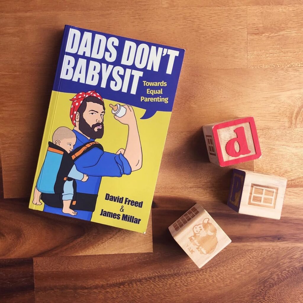 Dad's Don't Babysit book shows a Dad in the pose of Rosie the Riveter with a bottle in one hand and a baby on his back. Three wooden alphabet blocks sit beside the book.