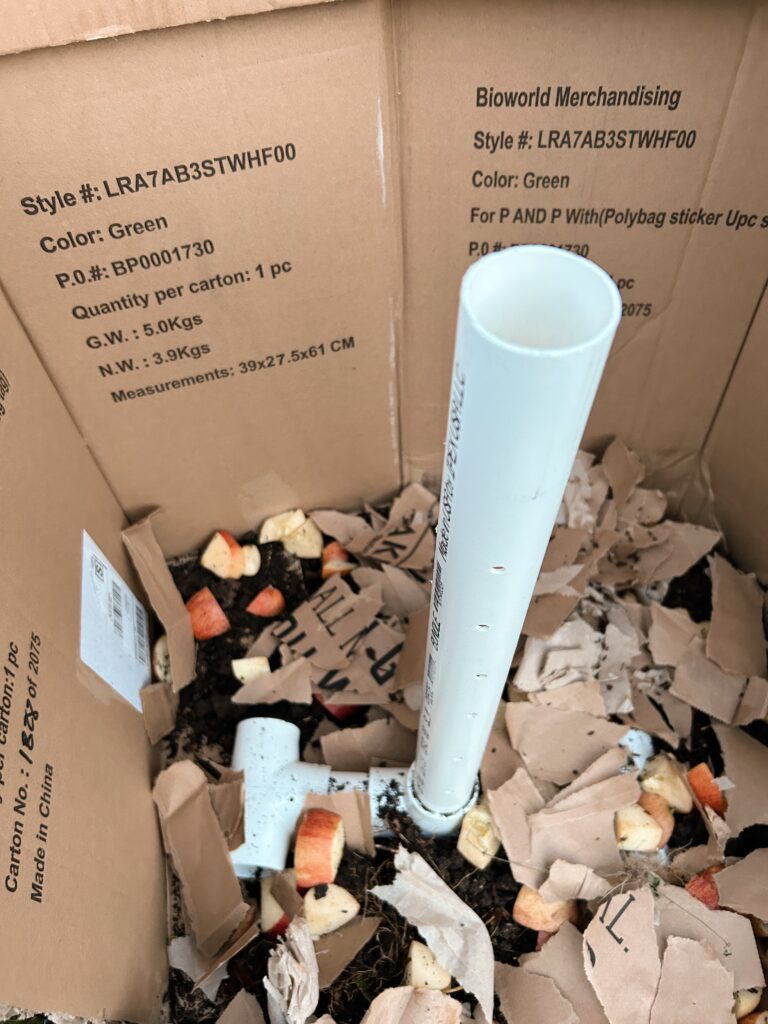 Compost heap with apples, cardboard and a white PVC pipes with holes in it set up vetyically in thr cnterz
