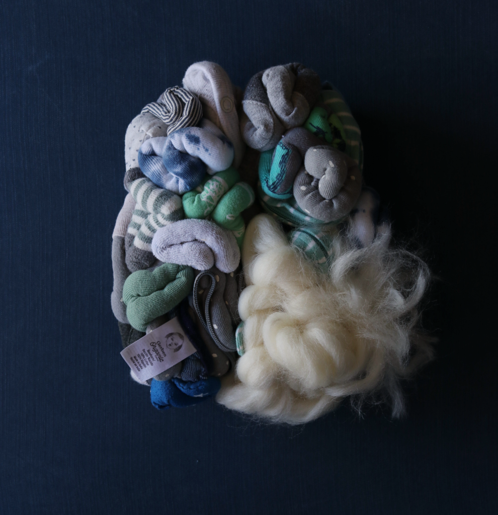 Close up of soft sculpture of a brain made from baby socks, onesies and washclothes. The lower right hemisphere is made from tangled wool roving which is tufting outward. It is centered on a square panel covered in navy cloth.