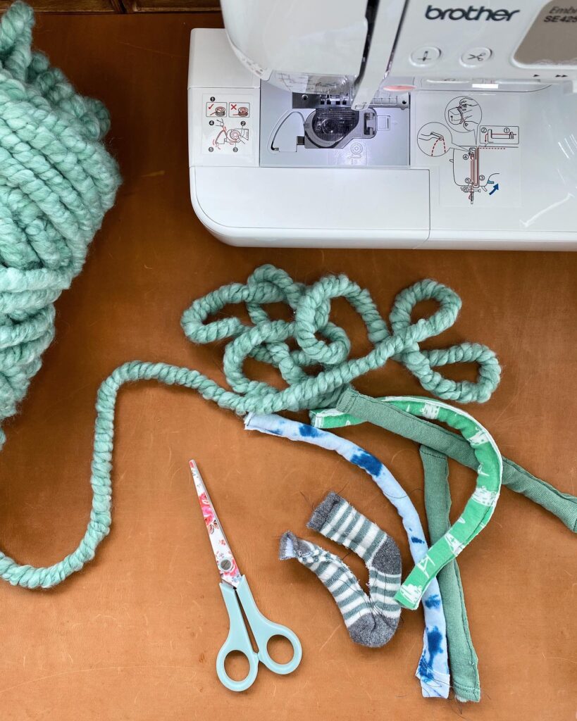 Workspace with sewing machine, scissors, chunky yard, and a baby sock which has been cut in half lengthwise.