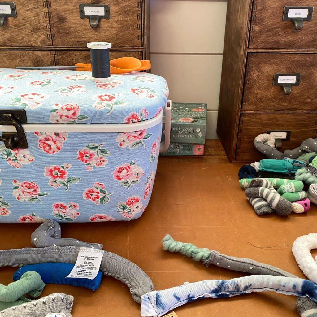 Blue sewing kit on a desk covered with snippets of chunky yarn encased in scraps of baby clothes.