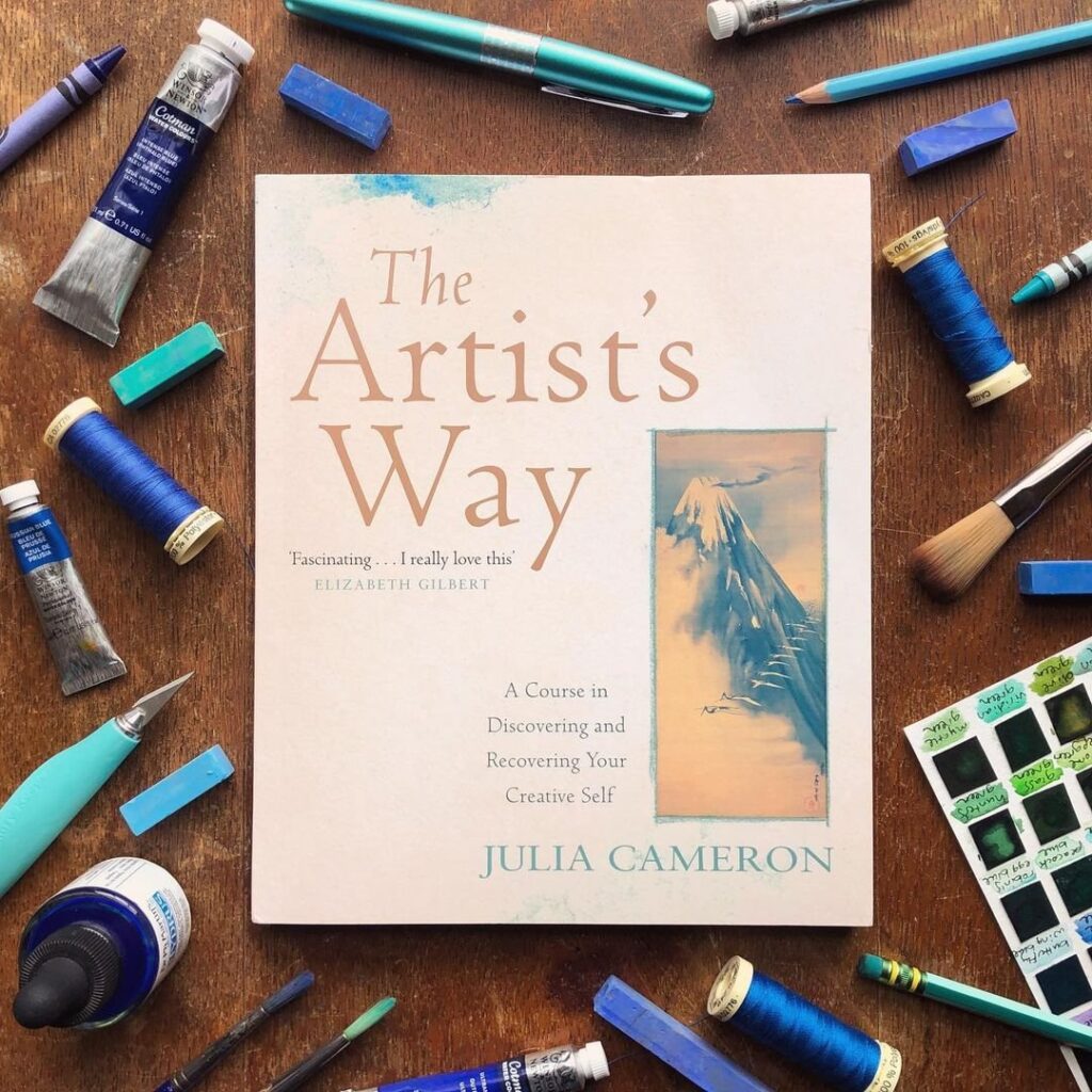 UK edition of The Artist's Way by Julia Cameron surrounded by blue art supplies: paints, pencils, pastels, thread and and brushes.