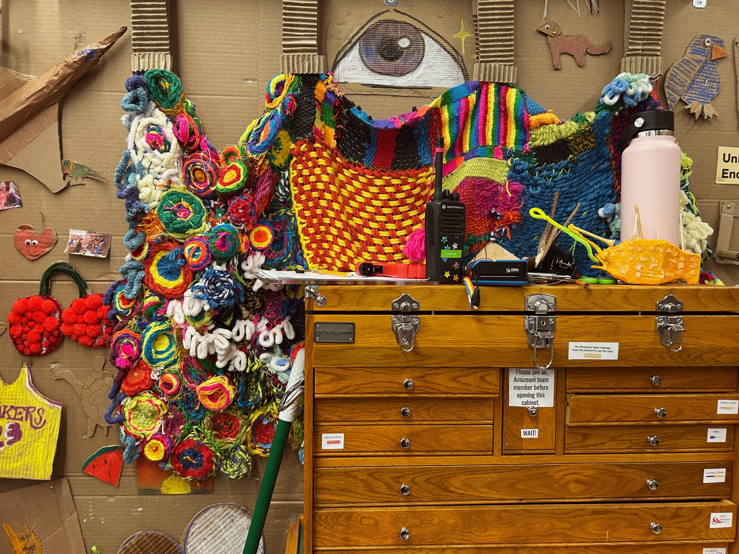 Maker space at children's museum a cardboard wall covered in art projects including a rainbow weaving an a giant eye. A wooden cabinet with creative tools is in front.