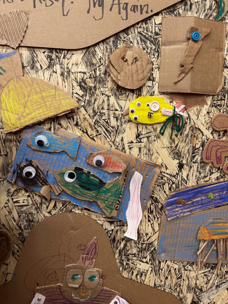 Particle board wall at science museum covered in cardboard art including fish with google eyes
