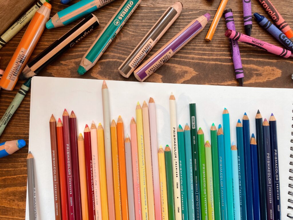 Rainbow of colored pencils on top of a sketchbook surrounded by art supplies